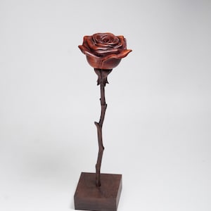 May include: A handcrafted wooden rose sculpture. The rose is a rich, reddish-brown color, with detailed petals. It sits atop a slender, dark brown stem, mounted on a square base. The sculpture is a decorative item.
