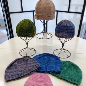 May include: A selection of hand-knitted beanies in shades of green, blue, and pink. Several beanies are displayed on black wire hat stands. The hats feature a ribbed brim and a simple, classic design.