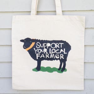 May include: A white canvas tote bag with black handles. The bag has a black and white illustration of a sheep with the text "Support Your Local Farmer" printed on it.