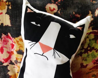 May include: Black and white cat pillow with a pink nose. The pillow is on a floral patterned fabric.