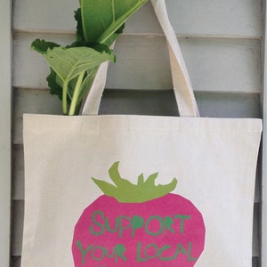 May include: A white canvas tote bag with a pink strawberry design. The strawberry has green leaves and the words "Support Your Local Farmer" written in green on it.