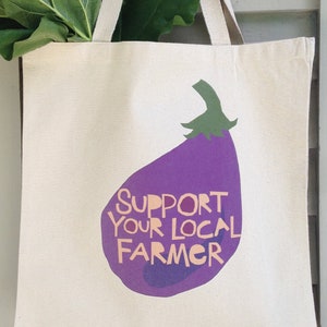 May include: A white canvas tote bag with a purple eggplant graphic and the text "SUPPORT YOUR LOCAL FARMER".