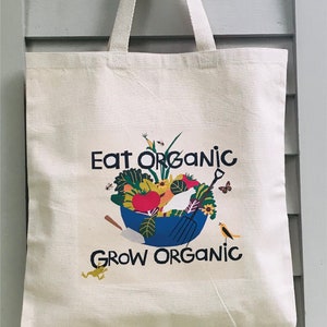 May include: A white canvas tote bag with a blue bowl filled with colorful vegetables, a white bird, and a yellow bird. The text "Eat Organic" is above the bowl and "Grow Organic" is below the bowl.