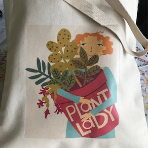 May include: Cream-colored tote bag with a graphic of a woman holding a red flower pot filled with plants. The words "PLANT LADY" are printed on the pot. The bag has a beige strap and is filled with greenery.