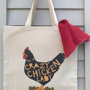 May include: A white canvas tote bag with black and yellow text that reads "Not So Crazy Chicken Lady" and a black silhouette of a chicken with the text incorporated into the design. The bag has red fabric draped over the top.