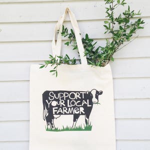 May include: A white canvas tote bag with black handles and a black and white cow graphic. The cow is eating grass and the text "SUPPORTS YOUR LOCAL FARMER" is printed above the cow.