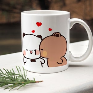 Peut inclure: Une tasse en céramique blanche avec une illustration de deux personnages de dessin animé, un panda et un ours brun, avec des cœurs rouges. Le panda porte un nœud papillon noir et l'ours embrasse le panda sur la joue.