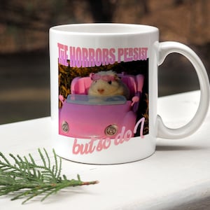 May include: White ceramic mug with a pink car and a hamster wearing sunglasses. The text on the mug reads "The horrors persist but so do I."