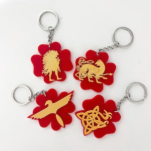 May include: Four red clover-shaped keychains with gold-colored animal and mythical creature designs. The designs include a bird, a wolf, a human figure, and a knot.
