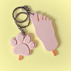 Könnte beinhalten: Ein rosa Pfotenabdruck und ein rosa fußförmiger Schlüsselanhänger mit einem silbernen Schlüsselring. Beide Anhänger haben einen Eis am Stiel befestigt.