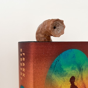 May include: A brown wooden bookend shaped like a curved horn with a hole in the end. The bookend is on top of a book with a red, orange, yellow, green, and blue cover. The book has the text "FRANK HERBERT" printed on the spine.