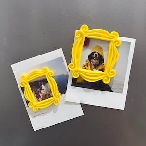 Peut inclure: Deux cadres en plastique jaune en forme du logo emblématique de l'émission "Friends". Le plus grand cadre est sur une photo d'un chien portant un imperméable jaune. Le plus petit cadre est sur une photo de deux personnes debout devant une plage.