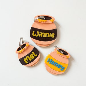 May include: Three honey pot-shaped keychains with different names on them. The keychains are orange with a brown band around the middle. The names are Winnie, Mel, and Honey.
