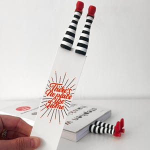 May include: A whimsical bookmark featuring black and white striped legs with red shoes. The bookmark has the text "There's No Place Like Home" in red. The legs are also used as a bookend.