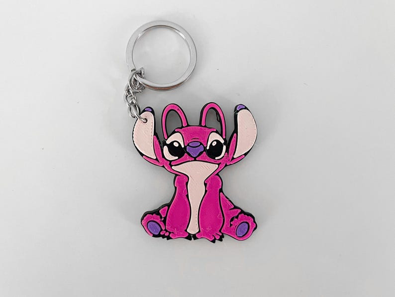 Pode incluir: Chaveiro Stitch rosa com um chaveiro prateado. O chaveiro apresenta um personagem de desenho animado Stitch com olhos grandes e um corpo rosa.