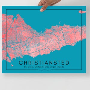 Op de afbeelding: Een turquoise kaartafdruk van Christiansted, St. Croix, Amerikaanse Maagdeneilanden. De kaart heeft een koraalkleurig topografisch ontwerp met blauwe lijnen en tekst. De afdruk wordt door een hand vastgehouden.
