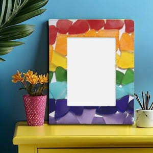 May include: A rectangular photo frame with a rainbow-colored mosaic design. The frame's border features a pattern of red, orange, yellow, green, blue, and purple glass-like pieces. The frame is on a yellow surface with a pink flower pot and a small white and gray container.