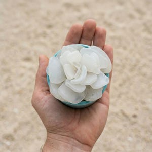 May include: A small, round, turquoise bowl filled with smooth, white sea glass pieces, held in an open hand. The sea glass is arranged to resemble a flower. The background is a sandy beach.