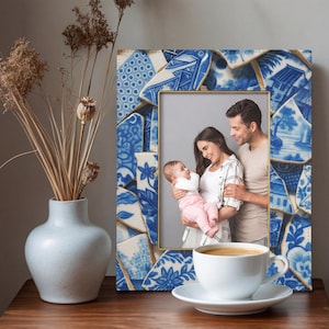 Puede incluir: Un marco de fotos de cerámica azul y blanca con borde dorado. El marco está decorado con trozos rotos de porcelana azul y blanca. Contiene una foto familiar. Una taza de café blanca está frente al marco.