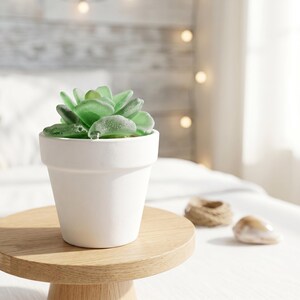 May include: A small, white ceramic pot holds a faux succulent with light green, textured leaves. The pot sits on a round, light-colored wooden stand. The background includes a bed and soft lighting.
