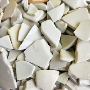 May include: A close-up shot of a pile of broken ceramic pieces. The fragments are primarily off-white with some variations in tone, and they have irregular shapes and sizes. The pieces appear to be from a larger object, possibly a plate or tile.