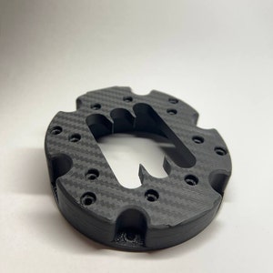 Könnte beinhalten: Ein schwarzes, 3D-gedrucktes Teil aus Kohlefaser mit einer runden Form und mehreren Löchern. Das Teil hat ein einzigartiges Design mit einem Ausschnitt in der Mitte.