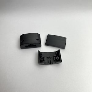 Op de afbeelding: Drie zwarte plastic componenten zijn gerangschikt op een witte ondergrond. De stukken omvatten een gebogen rechthoekig stuk, een plat rechthoekig stuk en een stuk met een gebogen interieur en twee gaten.