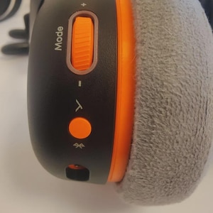 Può includere: Cuffie wireless nere e arancioni con un cuscinetto auricolare grigio e morbido. Le cuffie hanno un pulsante modalità e controlli del volume. Sono visibili anche un piccolo pulsante arancione e un simbolo Bluetooth.