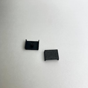 Könnte beinhalten: Zwei kleine schwarze Kunststoffkomponenten werden vor einem weißen Hintergrund gezeigt. Ein Teil hat eine rechteckige Form mit strukturierter Oberfläche und einer Lippe an jedem Ende. Das andere Teil hat eine rechteckige Basis mit einem kreisförmigen Loch und zwei vertikalen Verlängerungen.