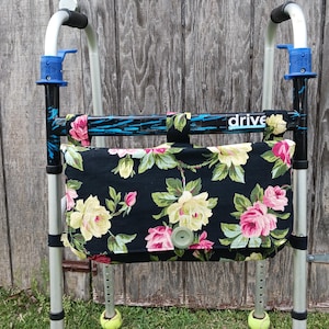 May include: A black fabric walker bag with a floral pattern of pink and yellow roses and green leaves. The bag is attached to a silver and black walker with the word "drive" printed on the front.