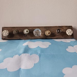 May include: A rustic wooden coat rack featuring seven decorative knobs in various styles. The knobs are made of materials like ceramic and glass, with colors including white, brown, and clear. The rack is mounted on a light blue surface with white cloud patterns.