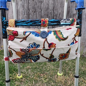 May include: A cream-colored walker bag with a vibrant floral pattern in red, blue, and green. The bag has a large blue button closure and is attached to a walker. The word "drive" is visible on the walker.
