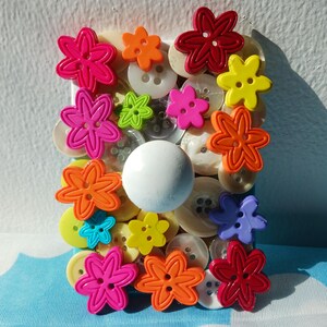 May include: A rectangular arrangement of various colorful flower-shaped buttons in shades of pink, orange, yellow, green, and red. The flowers are interspersed with a collection of white and yellow buttons, and a large white button.