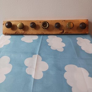May include: A wooden coat rack with seven decorative knobs. The knobs are various colors and designs, including bronze and gold tones. The wood has a natural finish with visible grain and knots. The rack is mounted on a light-colored wall.