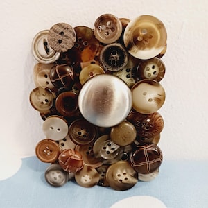 May include: A rectangular collage of various brown and tan buttons, including a large, silver-colored button in the center. The buttons vary in size, shape, and texture, creating a unique and tactile design. The background is a light blue surface.