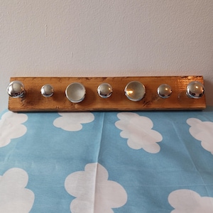 May include: A wooden coat rack with seven chrome knobs of varying sizes. The knobs are attached to a rectangular piece of stained wood. The rack is displayed against a white wall and a blue and white cloud-patterned fabric.