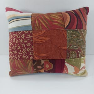 May include: A patchwork pillow with a variety of patterned fabrics in shades of red, orange, green, and cream. The pillow features a mix of floral, paisley, and geometric designs, creating a unique and textured appearance. The pillow is square-shaped.