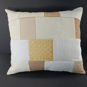 May include: A square decorative pillow with a patchwork design. The pillow features various textured fabrics in shades of cream, beige, and tan. The pillow has a soft, inviting appearance, perfect for adding a touch of comfort and style to any living space.