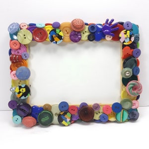 May include: A rectangular picture frame decorated with a variety of colorful buttons. The buttons are in shades of blue, red, yellow, green, and purple, and are of various sizes and shapes. The frame has a light wood border.