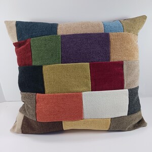 May include: Square patchwork pillow with a variety of colors including red, green, purple, and beige. The pillow has a textured fabric and a rectangular pattern. The pillow is a decorative home accessory.