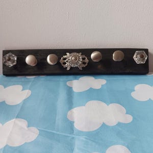 May include: A dark stained wooden coat rack with six decorative knobs. The knobs include clear faceted glass, silver-toned round knobs, and an ornate silver-toned center piece. The rack is mounted on a white wall.