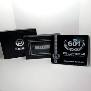 May include: Three black boxes with white text and logos. The box on the left has the word "PLASENC" and a logo. The middle box has the word "JAVA" and "Drew Estate". The box on the right has "601 BLACK" and "EXCLUSIVE ANNUAL RELEASE - 2021".
