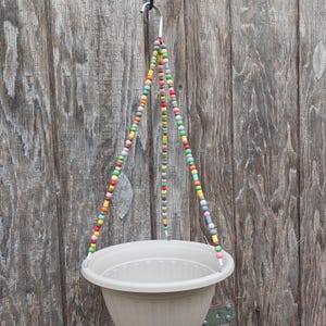 May include: A beige hanging planter with colorful beaded strands. The planter is suspended from a metal hook by three beaded strands, each featuring a variety of colors. The background is a weathered wooden fence.