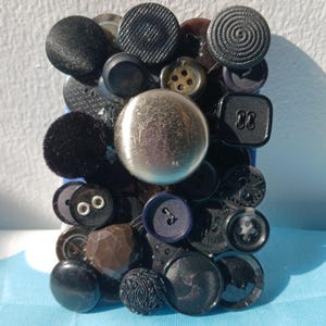 May include: A collection of various black buttons, including round, square, and textured designs, with a large silver button in the center. The buttons are arranged on a blue surface, creating a visually interesting display.