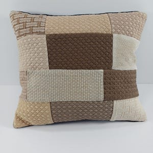 May include: Square decorative pillow with a patchwork design. The pillow features various textured fabrics in shades of brown, beige, and cream. The pillow has a black border and is likely used for home decor.