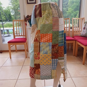 May include: A patchwork skirt displayed on a mannequin. The skirt is composed of various fabric squares in shades of blue, green, orange, and red. The skirt has a slit on the side. The skirt is a unique piece of clothing.