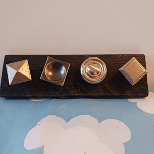 May include: A black wooden board with four decorative metal knobs of varying shapes: a pyramid, a square with a rounded center, a circular knob, and a square. The knobs are silver and metallic.