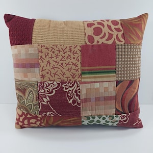 May include: A decorative patchwork pillow featuring a variety of fabrics in shades of burgundy, tan, and cream. The pillow has a rectangular shape and is composed of multiple fabric squares with different patterns and textures, creating a unique and cozy design.