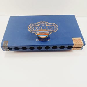 May include: A blue cigar box with the text "THE OSCAR" in gold lettering and a colorful, striped button. The box also has the word "CONNECTICUT" printed on it.