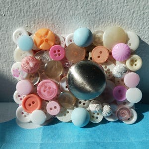 May include: A rectangular collage of various buttons in shades of white, pink, orange, and blue. A large, silver, spherical object is centered within the button arrangement. The buttons vary in size, shape, and texture, creating a unique decorative piece.